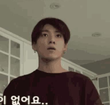 a young man is standing in a kitchen wearing a maroon shirt and looking at the camera .