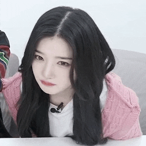 a woman with long black hair is sitting at a table wearing a pink sweater and looking at the camera .