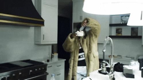 a man in a fur coat is standing in a kitchen drinking from a bottle ..