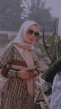 a woman in a hijab is standing in front of a greenhouse holding a flower .