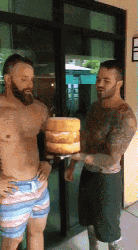 two shirtless men are standing next to each other and one is holding a large cake
