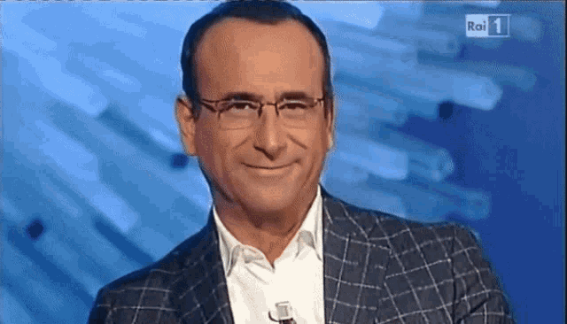 a man wearing glasses and a suit is smiling in front of a rai 1 screen