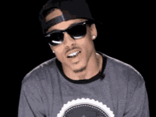 a man wearing sunglasses , a hat and a gray shirt is smiling .