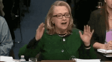 a woman in a green sweater and glasses is sitting at a table with her hands up .