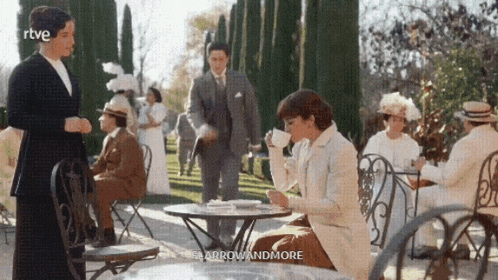 a woman is sitting at a table with a cup of tea in her hand while a man stands behind her ..
