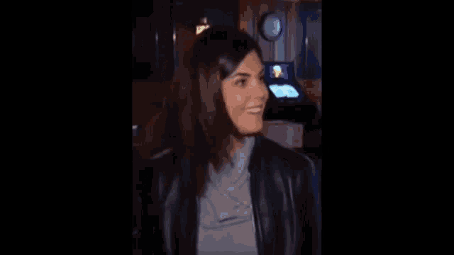 a woman in a leather jacket is smiling in a dark room with a clock on the wall .