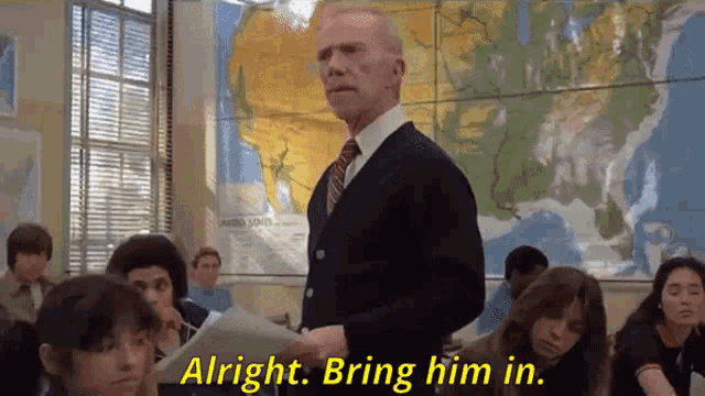 a man in a suit and tie stands in front of a group of students and says " alright bring him in "