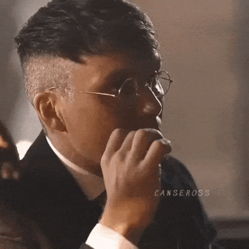 a man wearing glasses and a suit is sitting at a table with his hand on his chin .