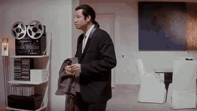 a man in a suit and tie is standing in a living room next to a shelf with tapes on it .