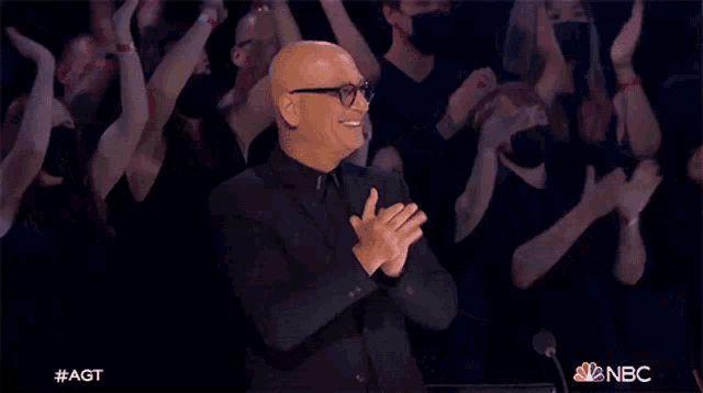 a man in a black suit and glasses is applauding in front of a crowd that is wearing masks