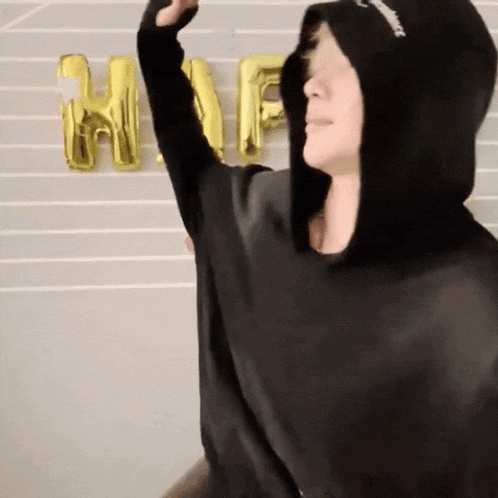 a person wearing a black hoodie is standing in front of a wall with balloons that spell out the word happy .