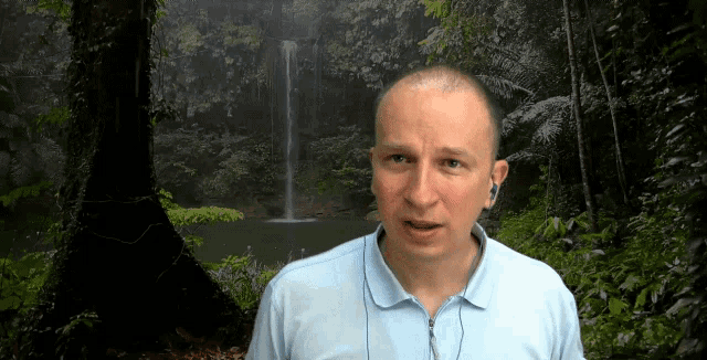 a man wearing ear buds stands in front of a waterfall in the woods