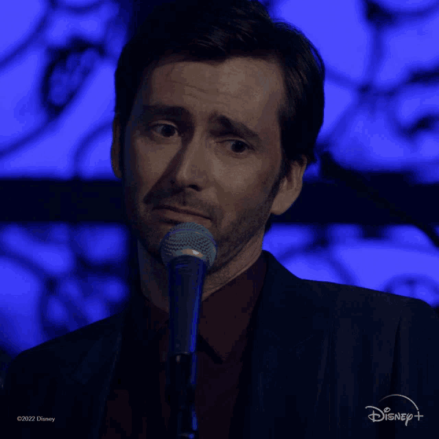 a man in a suit stands in front of a microphone with a disney logo in the background