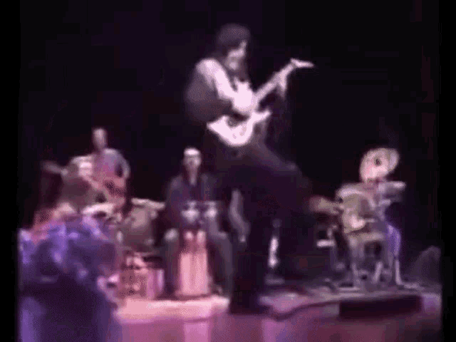 a man is playing a guitar on stage while a band plays drums .