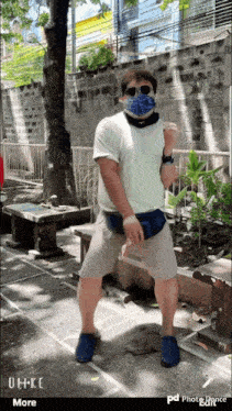 a man wearing a face mask and sunglasses stands in front of a brick wall and a tree