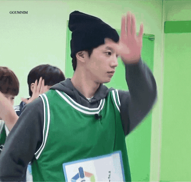 a young man wearing a green jersey and a black beanie is waving his hand in front of a green wall .