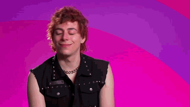 a man in a black vest is smiling with a picture of a woman behind him on a pink background .