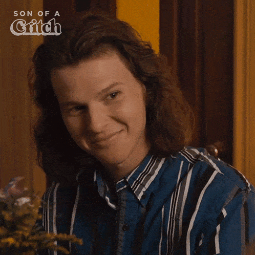 a man with long hair is smiling in front of a sign that says son of a witch