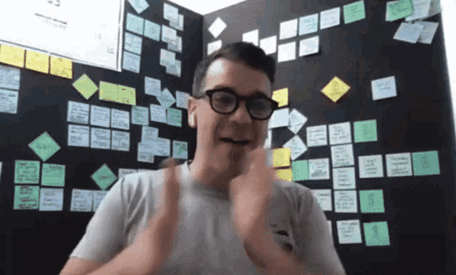a man wearing glasses is giving a thumbs up in front of a wall with post it notes on it