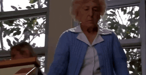 an elderly woman wearing a blue sweater and white shirt stands in front of a window
