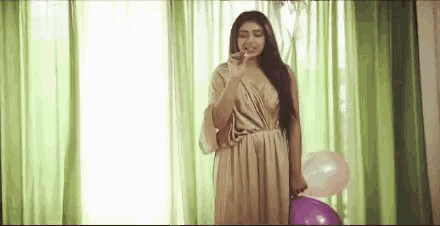 a woman in a long dress is standing in front of a window holding a purple balloon .