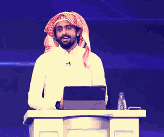 a man wearing a head scarf stands at a podium with a tablet