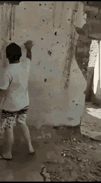 a man in a white shirt and shorts is standing in front of a wall with bullet holes in it .
