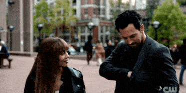 a man and a woman standing on a sidewalk with a cw logo on the bottom right