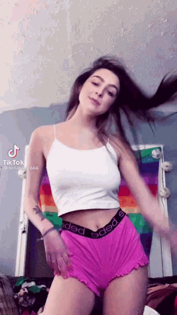 a woman wearing pink shorts and a white tank top is dancing in front of a rainbow flag .