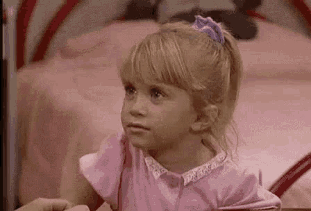 a little girl in a pink dress is sitting on a bed looking at the camera .