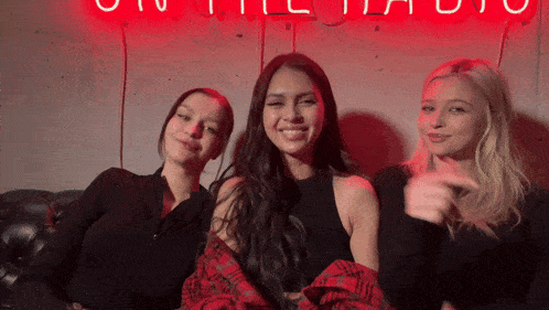 three women are posing for a picture in front of a neon sign that says ' u ' on it