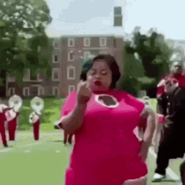 a woman in a pink dress is standing in front of a marching band and giving the middle finger .