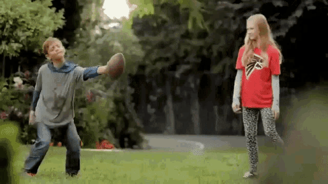 a boy and a girl are playing with a football in the grass . the boy is wearing a falcons shirt .