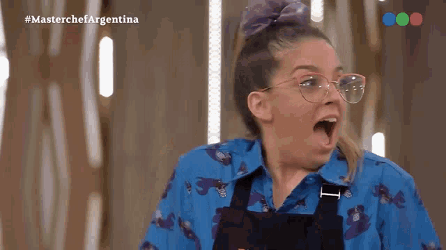 a woman wearing glasses and a blue shirt is making a surprised face while watching a masterchef argentina show