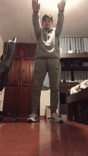 a young man wearing a grey hoodie and grey sweatpants stands in a bedroom with his arms in the air