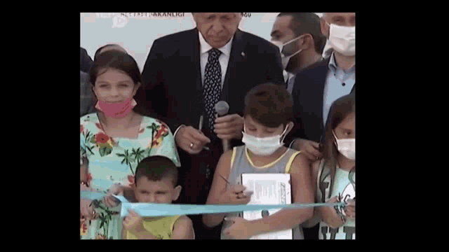 a man in a suit and tie is holding a microphone in front of a group of children wearing masks