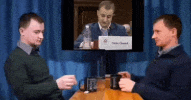 two men are sitting at a table talking to each other in front of a television screen .