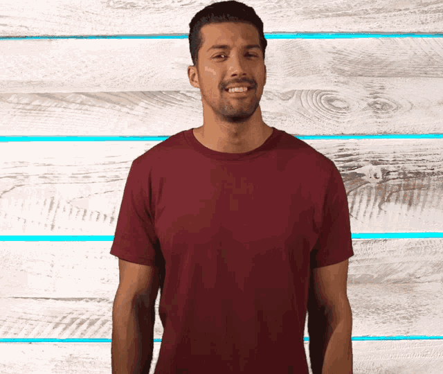 a man wearing a maroon t-shirt is standing in front of a wooden wall