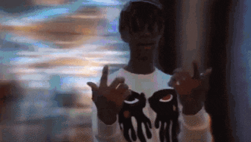 a man wearing a hat and a t-shirt with a picture of a hand on it is making a peace sign .