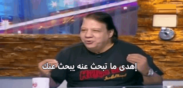 a man in a black shirt is sitting at a table with a cup of coffee and talking in arabic .