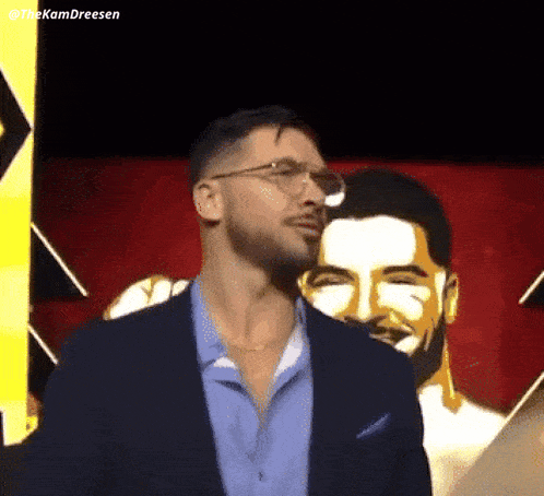 a man wearing glasses and a suit is standing in front of a painting of another man .