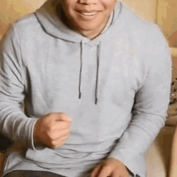 a man in a gray hoodie is sitting on a couch and smiling .