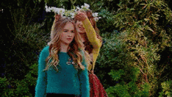 two girls are standing next to each other in a park . one of the girls is wearing a flower crown on her head .