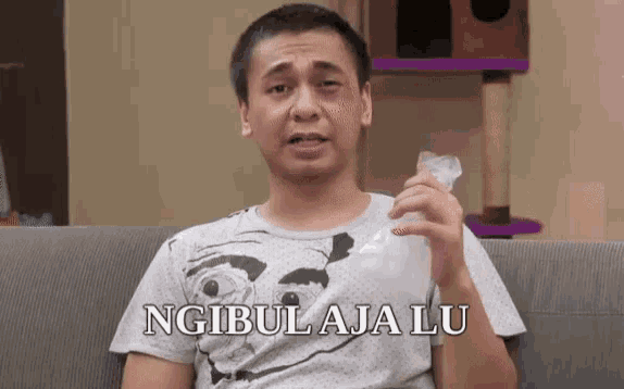a man is sitting on a couch holding a piece of paper with the words ngibu aja lu written on it .