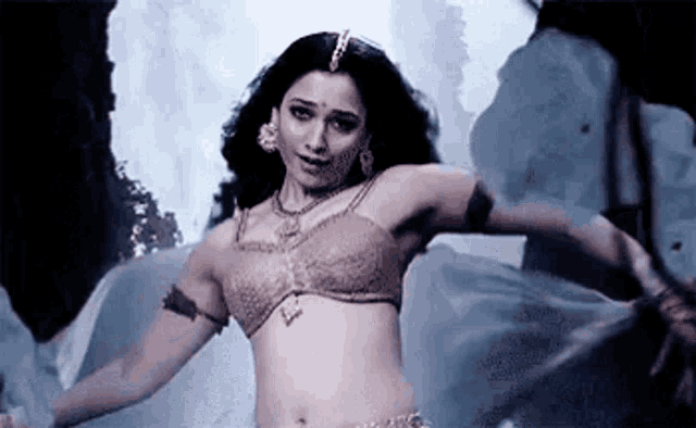 a woman is dancing in front of a waterfall while wearing a bra and earrings .