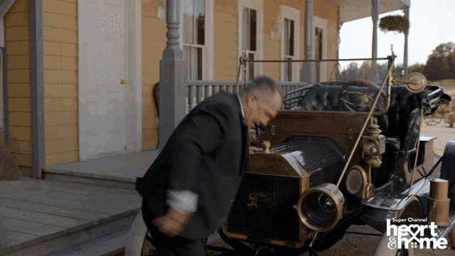 a man in a suit pushes a ford car in front of a yellow house