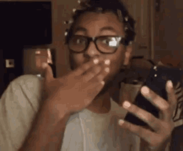 a man wearing glasses is covering his mouth with his hand while looking at his phone .