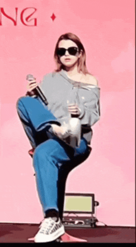 a woman wearing sunglasses is sitting on a chair holding a microphone in front of a pink background that says ng