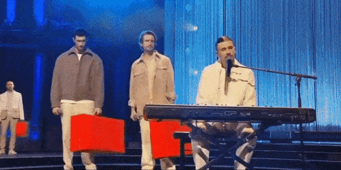 a man is singing into a microphone while a group of men stand behind him holding red boxes