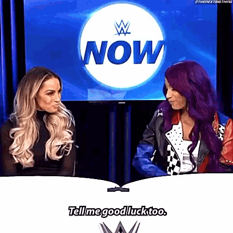 two women are sitting at a table and talking to each other in front of a wrestling show .
